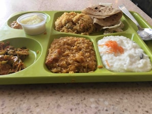 A green segmented tray contains a variety of Indian dishes including rice, flatbread, a lentil dish, yogurt garnished with grated carrot, and vegetables. A small container with a creamy dessert is also visible.