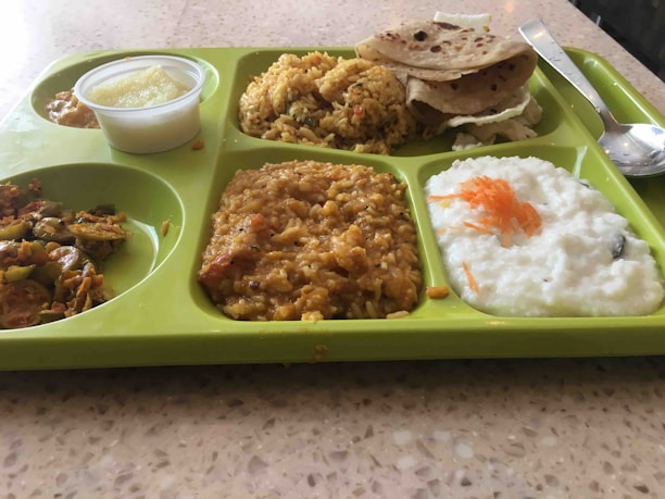 A green segmented tray contains a variety of Indian dishes including rice, flatbread, a lentil dish, yogurt garnished with grated carrot, and vegetables. A small container with a creamy dessert is also visible.