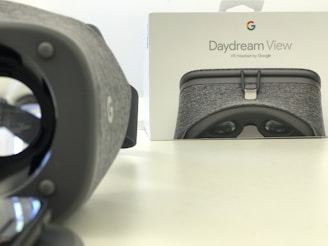 A Daydream View VR headset is positioned prominently on the left, with its gray fabric texture visible. In the background, there is a box featuring an image of the same headset and the Google logo. The setting is minimalist with a light-colored, smooth surface.