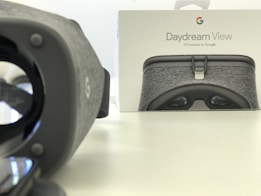A Daydream View VR headset is positioned prominently on the left, with its gray fabric texture visible. In the background, there is a box featuring an image of the same headset and the Google logo. The setting is minimalist with a light-colored, smooth surface.
