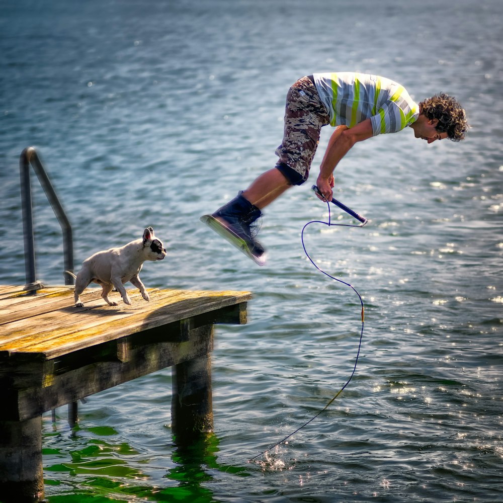 A man with curly hair diving into water, with his puppy on deck.