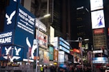 A bustling urban scene with numerous bright and colorful digital billboards displaying various advertisements, including ones for American Eagle and Coca-Cola. The area is crowded with people walking and some standing in groups, reflecting a lively atmosphere. Tall buildings surround the space, and it appears to be a famous urban location known for its vibrant lights and constant activity, likely during the evening.
