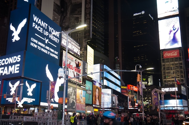 A bustling urban scene with numerous bright and colorful digital billboards displaying various advertisements, including ones for American Eagle and Coca-Cola. The area is crowded with people walking and some standing in groups, reflecting a lively atmosphere. Tall buildings surround the space, and it appears to be a famous urban location known for its vibrant lights and constant activity, likely during the evening.