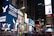 A bustling urban scene with numerous bright and colorful digital billboards displaying various advertisements, including ones for American Eagle and Coca-Cola. The area is crowded with people walking and some standing in groups, reflecting a lively atmosphere. Tall buildings surround the space, and it appears to be a famous urban location known for its vibrant lights and constant activity, likely during the evening.