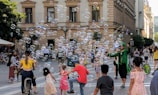 A lively street scene with people enjoying the playful presence of bubbles. A performer dressed in green is entertaining a crowd that includes children and adults. The setting is an urban area with historic architecture. People are casually dressed, indicating a relaxed atmosphere.