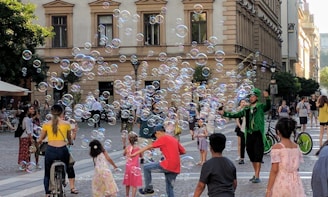 A lively street scene with people enjoying the playful presence of bubbles. A performer dressed in green is entertaining a crowd that includes children and adults. The setting is an urban area with historic architecture. People are casually dressed, indicating a relaxed atmosphere.