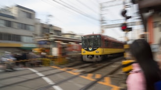 A train is moving quickly through an urban area, creating a sense of speed and motion. The surroundings, including buildings and a railroad crossing barrier, appear blurred, emphasizing the train's velocity. A person is partially visible in the foreground, also slightly blurred due to motion.