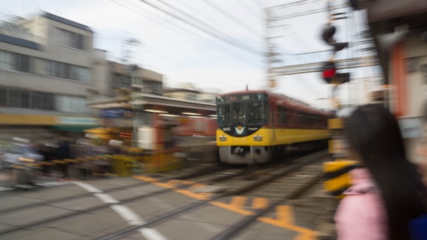 A train is moving quickly through an urban area, creating a sense of speed and motion. The surroundings, including buildings and a railroad crossing barrier, appear blurred, emphasizing the train's velocity. A person is partially visible in the foreground, also slightly blurred due to motion.