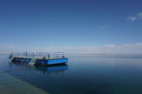 A large, flat blue barge with safety railings sits on calm water under a clear blue sky. The water reflects the barge and sky, creating a serene and peaceful scene. In the distance, a narrow strip of land is visible on the horizon, with a few clouds scattered across the sky.