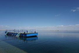 A large, flat blue barge with safety railings sits on calm water under a clear blue sky. The water reflects the barge and sky, creating a serene and peaceful scene. In the distance, a narrow strip of land is visible on the horizon, with a few clouds scattered across the sky.