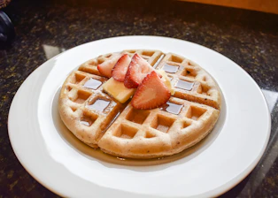 Close-up of a freshly made waffle topped with colorful fruits and melting chocolate syrup on a rustic wooden table.