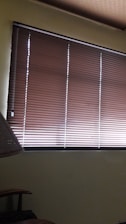 Brown horizontal blinds cover the window in a dimly lit room. The shades are partially closed, allowing minimal light to filter through. A portion of a chair or furniture piece can be seen in the bottom left corner.