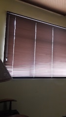 Brown horizontal blinds cover the window in a dimly lit room. The shades are partially closed, allowing minimal light to filter through. A portion of a chair or furniture piece can be seen in the bottom left corner.