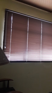Brown horizontal blinds cover the window in a dimly lit room. The shades are partially closed, allowing minimal light to filter through. A portion of a chair or furniture piece can be seen in the bottom left corner.