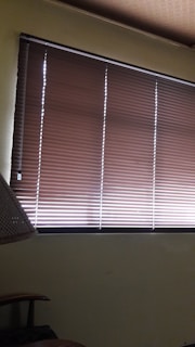 Brown horizontal blinds cover the window in a dimly lit room. The shades are partially closed, allowing minimal light to filter through. A portion of a chair or furniture piece can be seen in the bottom left corner.