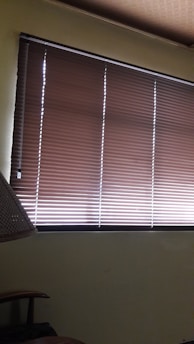Brown horizontal blinds cover the window in a dimly lit room. The shades are partially closed, allowing minimal light to filter through. A portion of a chair or furniture piece can be seen in the bottom left corner.