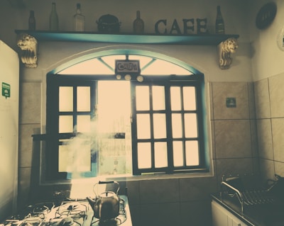 A cozy kitchen with a vintage look, featuring a window with grid-like panes allowing sunlight to stream in. Above the window are decorative elements, including bottles and the word 'CAFE'. A kettle is on the stove with steam rising, and there are tile walls and countertops.