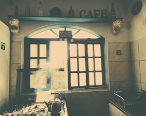 A cozy kitchen with a vintage look, featuring a window with grid-like panes allowing sunlight to stream in. Above the window are decorative elements, including bottles and the word 'CAFE'. A kettle is on the stove with steam rising, and there are tile walls and countertops.