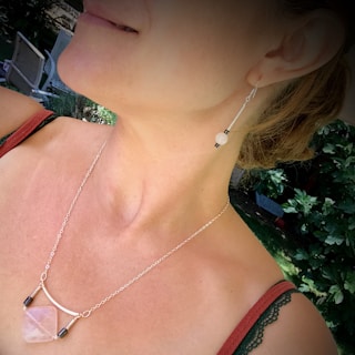 A close-up view of a woman's neck and part of her face, showcasing a dangling earring and a pendant necklace. The earring features a small bead, while the necklace has a geometric pendant. The background is blurry but filled with greenery, suggesting an outdoor setting.