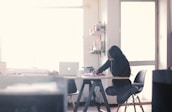 A person with long hair sits at a desk in a bright room, focusing intently on writing or drawing. The room is softly lit with natural light coming from large windows. There are shelves mounted on the wall with books and objects, and a laptop is open on the desk in front of them.