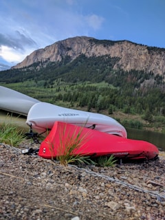 Two kayaks, one red and one white, are resting on a pebbled shore beside a calm body of water. In the background, a lush green forest blankets the lower slopes of a rocky mountain, and the sky is partly cloudy with patches of blue.