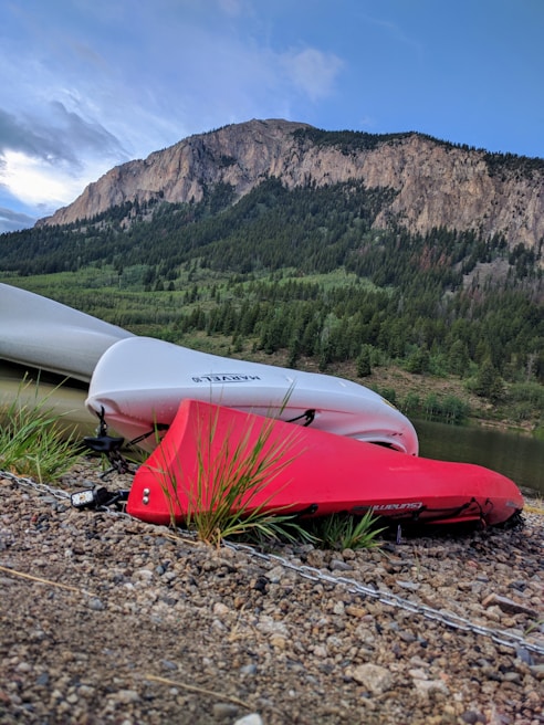 Two kayaks, one red and one white, are resting on a pebbled shore beside a calm body of water. In the background, a lush green forest blankets the lower slopes of a rocky mountain, and the sky is partly cloudy with patches of blue.