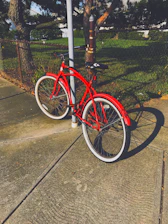 A bright red kids' bike leaning against a sunny park bench with colorful balloons tied to the handlebars.