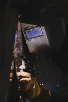 A person is welding a metal surface while wearing a protective welding helmet and glove. Bright sparks and faint smoke are visible as the welding rod makes contact with the metal.