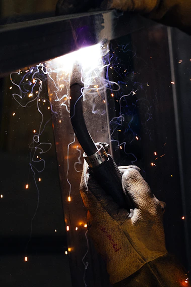 Close-up of a skilled welder's hands steadying a glowing welding torch, sparks flying gently in a dim workshop.