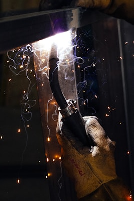 A gloved hand firmly grips a welding torch, emitting bright sparks and swirls of smoke. The intense light from the welding process illuminates the scene, creating a contrast against the dimly lit background.