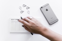 A hand is holding a pen over a weekly planner with sections for different days, placed on a white surface. Nearby, there is a silver iPhone and several metal paper clips.