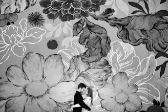 A couple embracing in front of a large mural featuring oversized flowers and leaves in various shades of black, white, and gray. The artwork is stylized with bold outlines and intricate patterns, creating a dramatic and romantic backdrop.