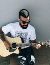 A man with short hair and a beard is seated, playing an acoustic guitar. He is wearing sunglasses, a white t-shirt with a graphic design, and dark pants. The setting appears to be outdoors with a tiled floor and a plain background.