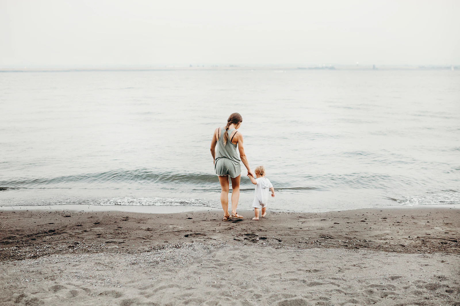 A mother and baby walking along a pale sand seashore during daytime, evoking a family beach day