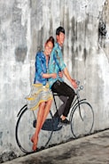 A mural on a textured, weathered wall depicts a joyful couple riding a bicycle. They appear lively, with the woman dressed in a striped blue and white skirt and a blue jacket, while the man wears a blue shirt and dark pants. The mural artist cleverly incorporates the real bicycle seat and handlebars into the painting, creating a seamless blend between reality and art.