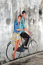 A mural on a textured, weathered wall depicts a joyful couple riding a bicycle. They appear lively, with the woman dressed in a striped blue and white skirt and a blue jacket, while the man wears a blue shirt and dark pants. The mural artist cleverly incorporates the real bicycle seat and handlebars into the painting, creating a seamless blend between reality and art.