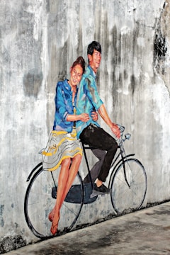 A mural on a textured, weathered wall depicts a joyful couple riding a bicycle. They appear lively, with the woman dressed in a striped blue and white skirt and a blue jacket, while the man wears a blue shirt and dark pants. The mural artist cleverly incorporates the real bicycle seat and handlebars into the painting, creating a seamless blend between reality and art.