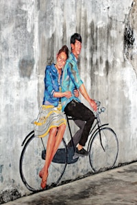 A mural on a textured, weathered wall depicts a joyful couple riding a bicycle. They appear lively, with the woman dressed in a striped blue and white skirt and a blue jacket, while the man wears a blue shirt and dark pants. The mural artist cleverly incorporates the real bicycle seat and handlebars into the painting, creating a seamless blend between reality and art.