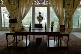 A beautifully decorated room with an ornate wooden desk in the foreground, adorned with decorative objects including a bust, a triangular sculpture, and a vase. Behind the desk are tall windows framed by sheer, elegant curtains, allowing natural light to illuminate the space. The room features intricate patterns on the walls and a richly colored Persian rug on the floor, contributing to the luxurious ambiance.