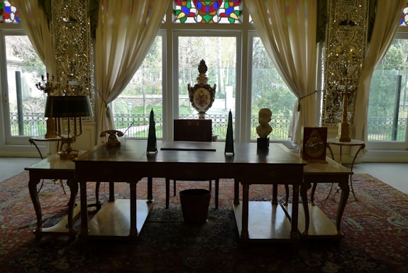 A beautifully decorated room with an ornate wooden desk in the foreground, adorned with decorative objects including a bust, a triangular sculpture, and a vase. Behind the desk are tall windows framed by sheer, elegant curtains, allowing natural light to illuminate the space. The room features intricate patterns on the walls and a richly colored Persian rug on the floor, contributing to the luxurious ambiance.
