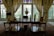 A beautifully decorated room with an ornate wooden desk in the foreground, adorned with decorative objects including a bust, a triangular sculpture, and a vase. Behind the desk are tall windows framed by sheer, elegant curtains, allowing natural light to illuminate the space. The room features intricate patterns on the walls and a richly colored Persian rug on the floor, contributing to the luxurious ambiance.