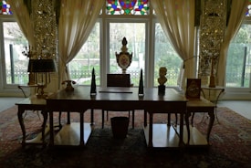 A beautifully decorated room with an ornate wooden desk in the foreground, adorned with decorative objects including a bust, a triangular sculpture, and a vase. Behind the desk are tall windows framed by sheer, elegant curtains, allowing natural light to illuminate the space. The room features intricate patterns on the walls and a richly colored Persian rug on the floor, contributing to the luxurious ambiance.