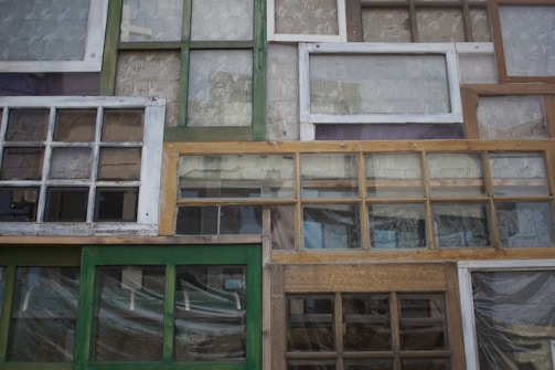 A collection of variously sized and styled window frames arranged in a mosaic-like pattern. The frames vary in color from green to white and brown, displaying a mix of vintage and rustic designs. Some have panes of glass, while others appear more weathered.