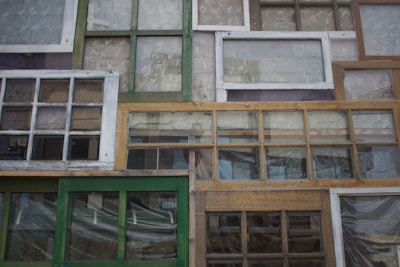 A collection of variously sized and styled window frames arranged in a mosaic-like pattern. The frames vary in color from green to white and brown, displaying a mix of vintage and rustic designs. Some have panes of glass, while others appear more weathered.