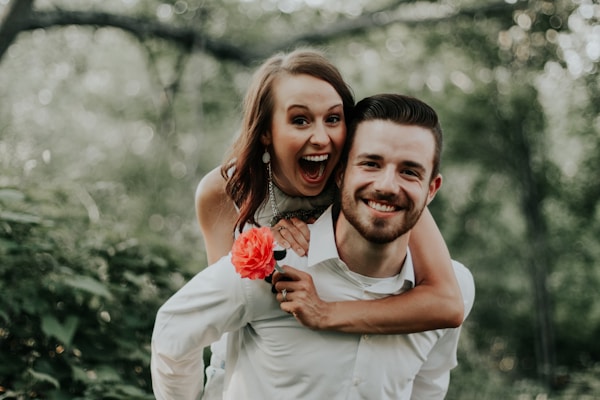 A smiling woman playfully wraps her arms around a man from behind in an outdoor setting. The man is also smiling, and the woman is holding a bright red flower. The background features lush green foliage, suggesting a forest or garden.
