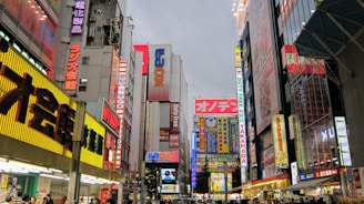 A bustling urban street scene dominated by tall buildings with numerous colorful neon signs and billboards. The signs display a mix of Japanese characters and English words, adding to the vibrant atmosphere. The street level is busy with people walking, indicating a lively and active commercial area.