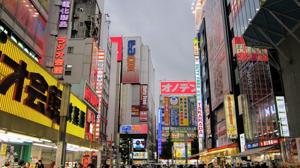 A bustling urban street scene dominated by tall buildings with numerous colorful neon signs and billboards. The signs display a mix of Japanese characters and English words, adding to the vibrant atmosphere. The street level is busy with people walking, indicating a lively and active commercial area.