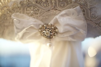 A detailed close-up of a decorative fabric featuring intricate lace patterns and embellishments. At the center, a large bow made of delicate white material is adorned with a sparkly brooch decorated with pearls and crystals.