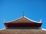 A traditional architecture style roof with a steep triangular shape adorned with intricate figures at its corners. The roof appears to be made of small brown tiles, set against a bright and clear blue sky.