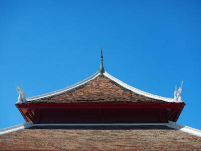 A traditional architecture style roof with a steep triangular shape adorned with intricate figures at its corners. The roof appears to be made of small brown tiles, set against a bright and clear blue sky.